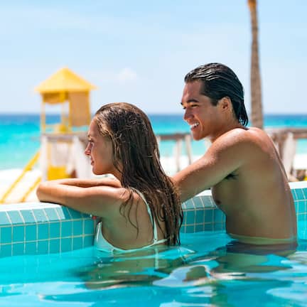 A couple in a swimming pool with a beach and a lifeguard tower in the background.