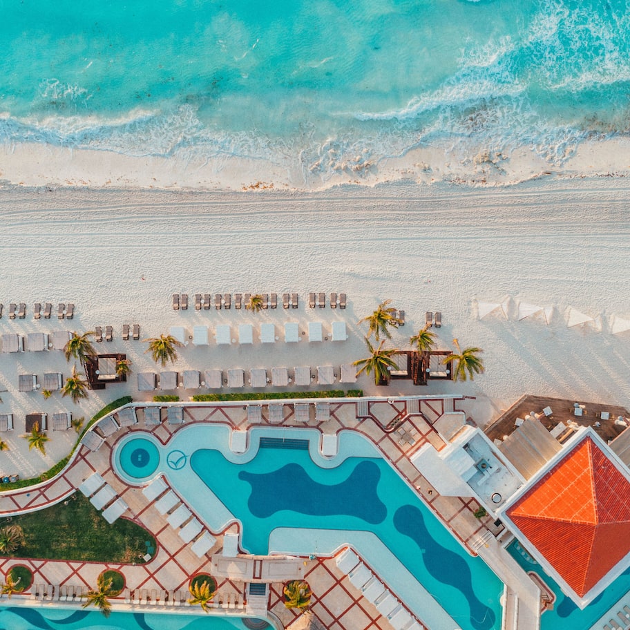 An aerial view of a resort with a large swimming pool, multiple lounge areas, and a beach.