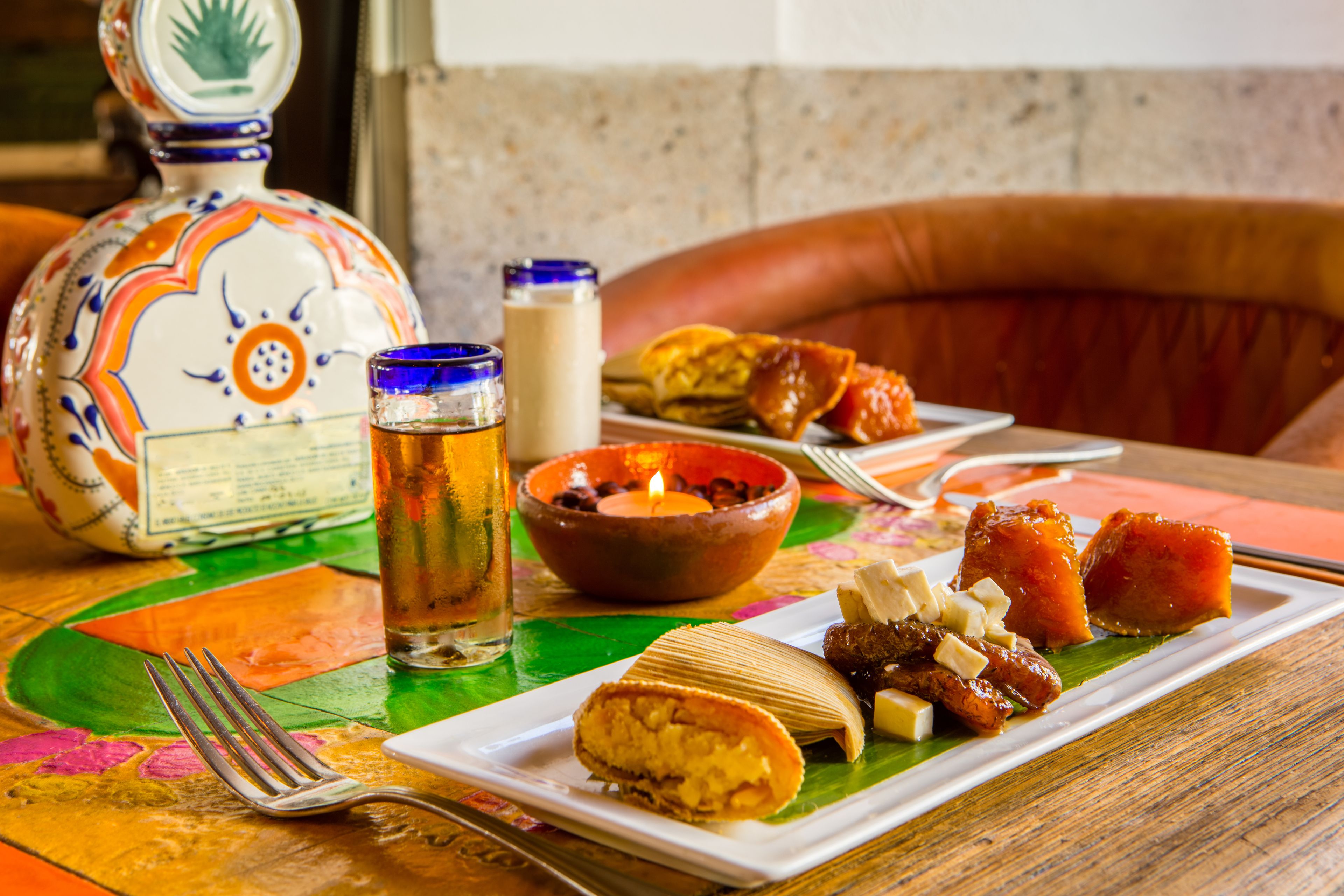 A table with a colorful tablecloth, a ceramic pitcher, a glass of iced tea, a candle, and a plate with various foods.