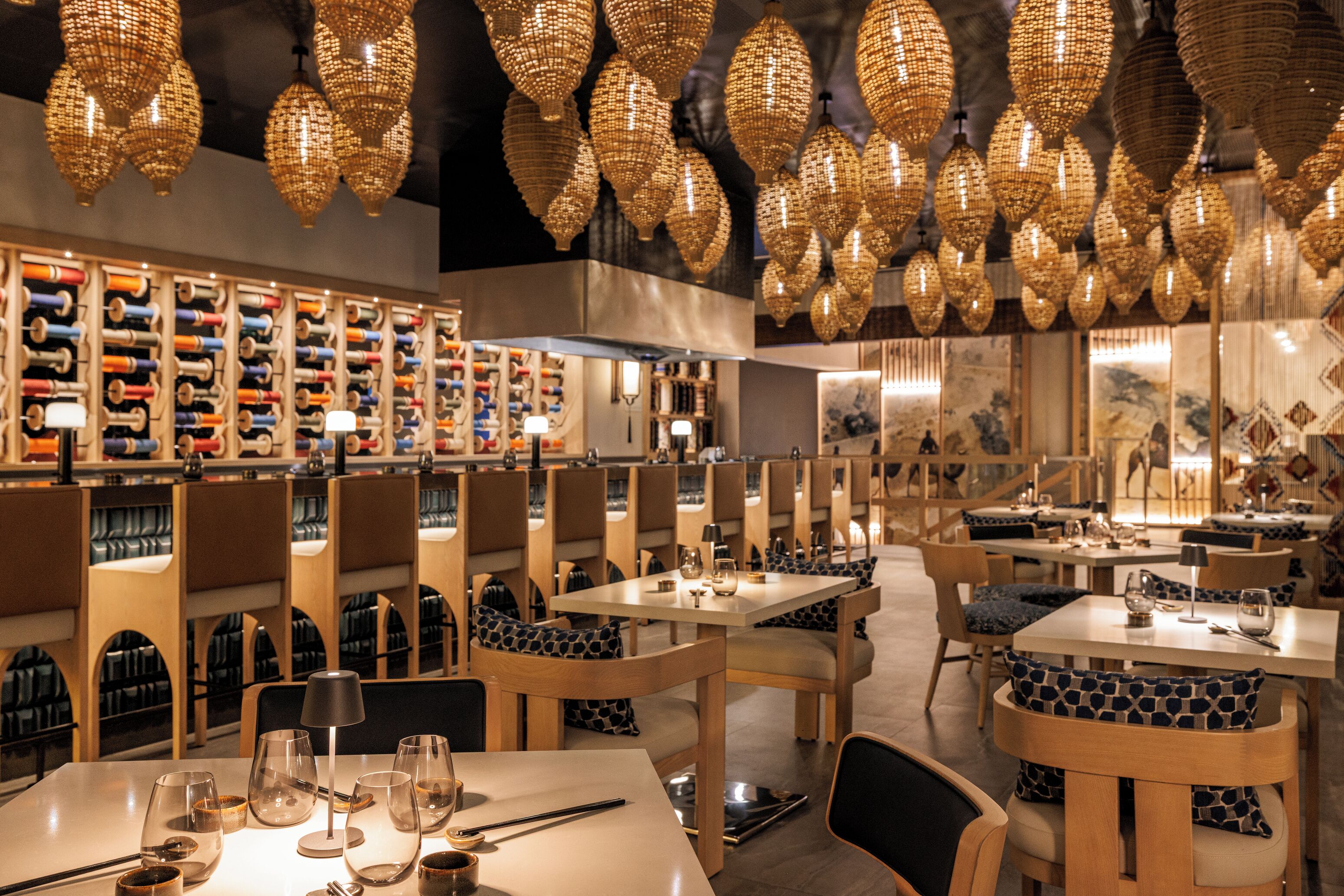 A restaurant with multiple tables and chairs, a wall of bottles, and hanging decorative lights.