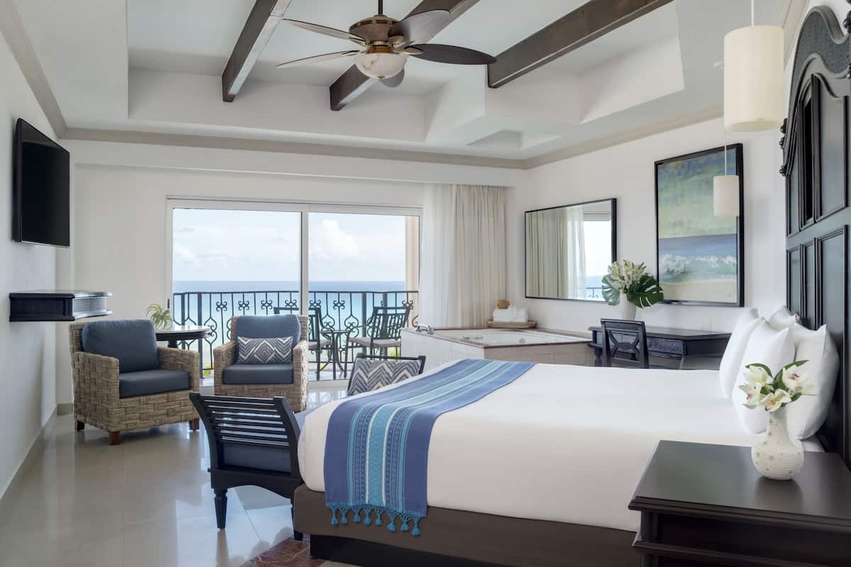 A hotel room with a large bed, a ceiling fan, a balcony with a view, wicker furniture, and a desk with a chair.