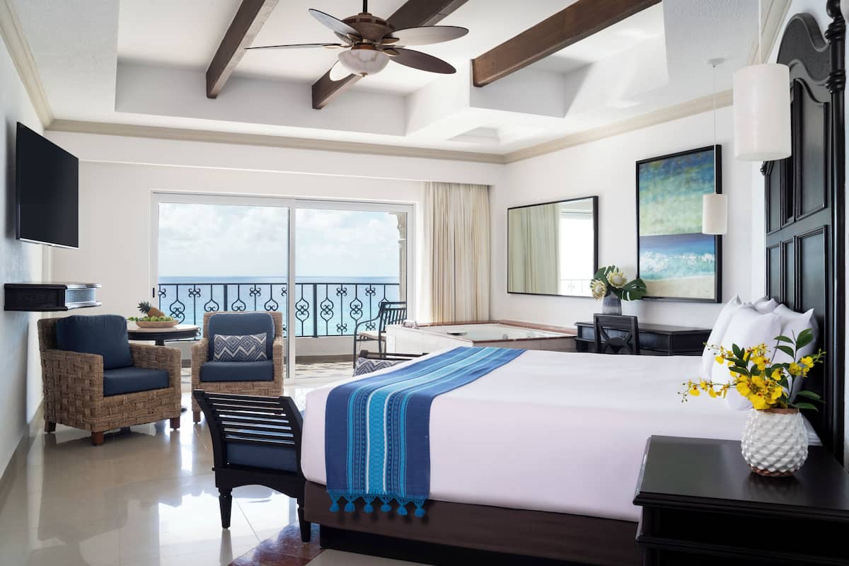 A hotel room with a large bed, a ceiling fan, a balcony with a view, wicker furniture, and a small table with a vase of flowers.