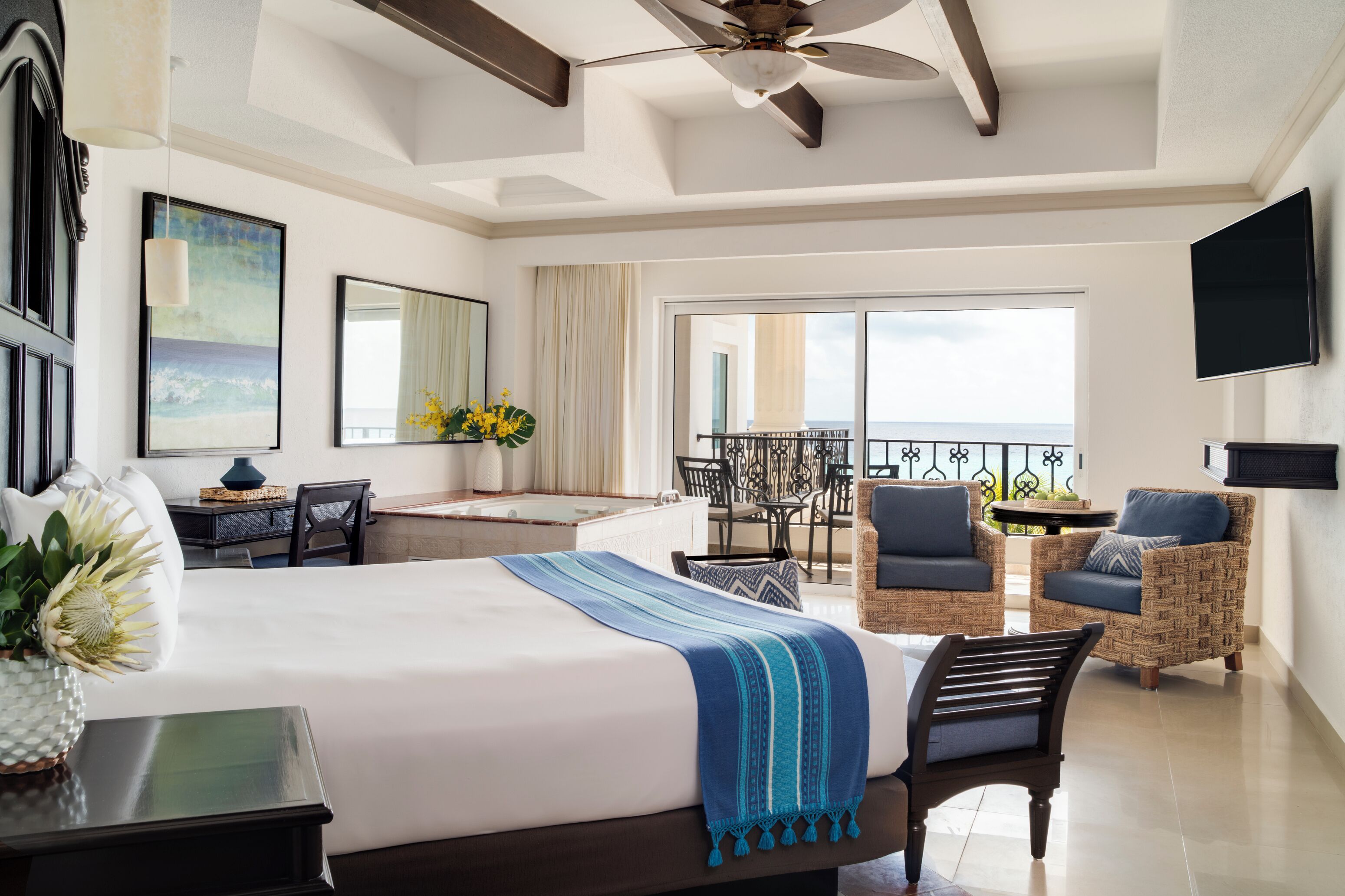A hotel room with a large bed, a desk, a chair, a balcony with a view, and a ceiling fan.