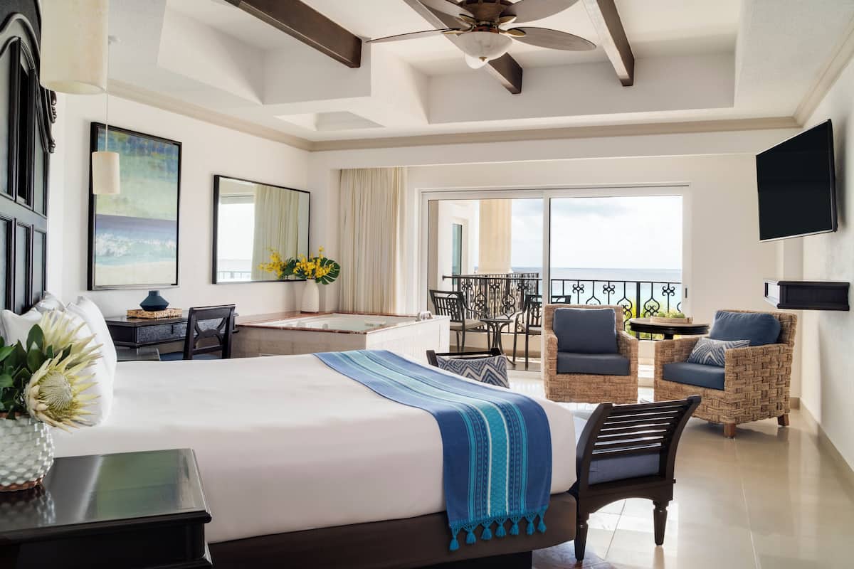 A hotel room with a large bed, a desk, a chair, a balcony with a view, and a ceiling fan.