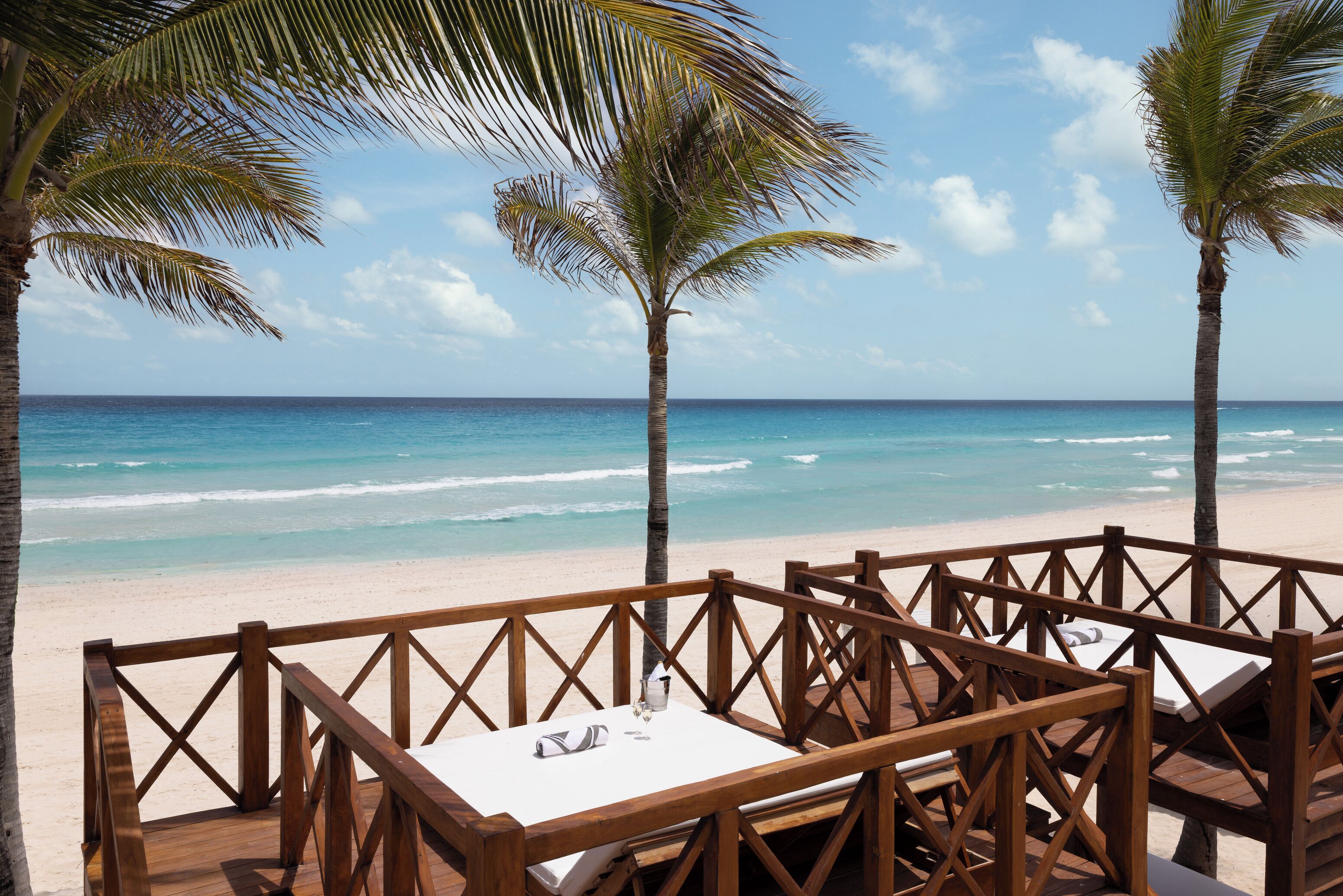 Una terraza de madera con una mesa y sillas que da a una playa con palmeras y el océano.