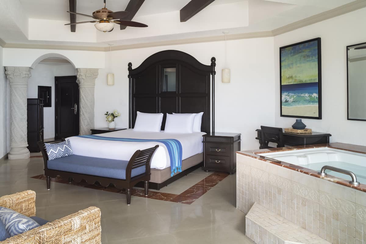A spacious bedroom with a large bed, a sitting area with a chair, a small table, a nightstand, a bathtub, and a ceiling fan.