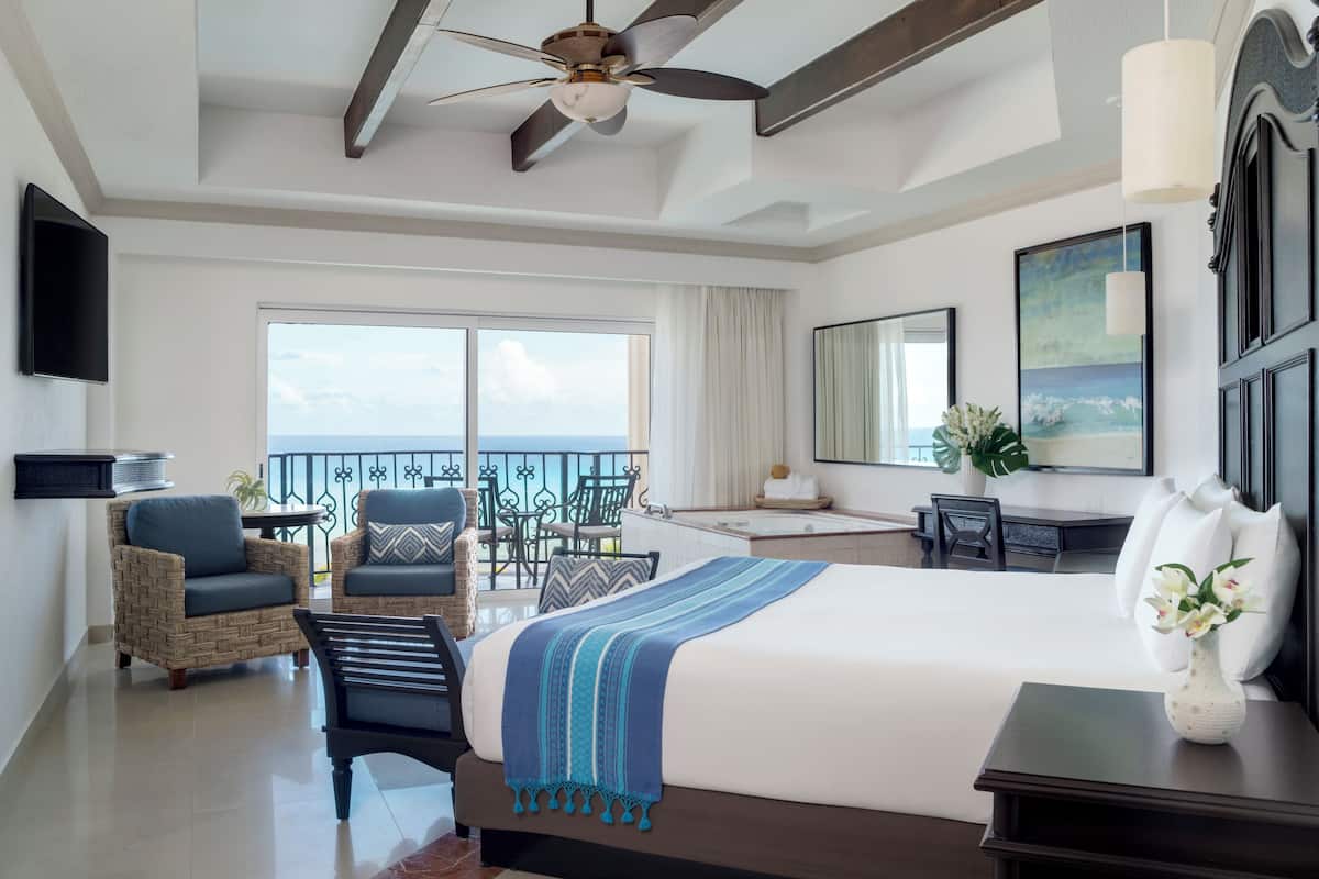 A hotel room with a bed, a desk, a chair, a balcony with a view, and a ceiling fan.