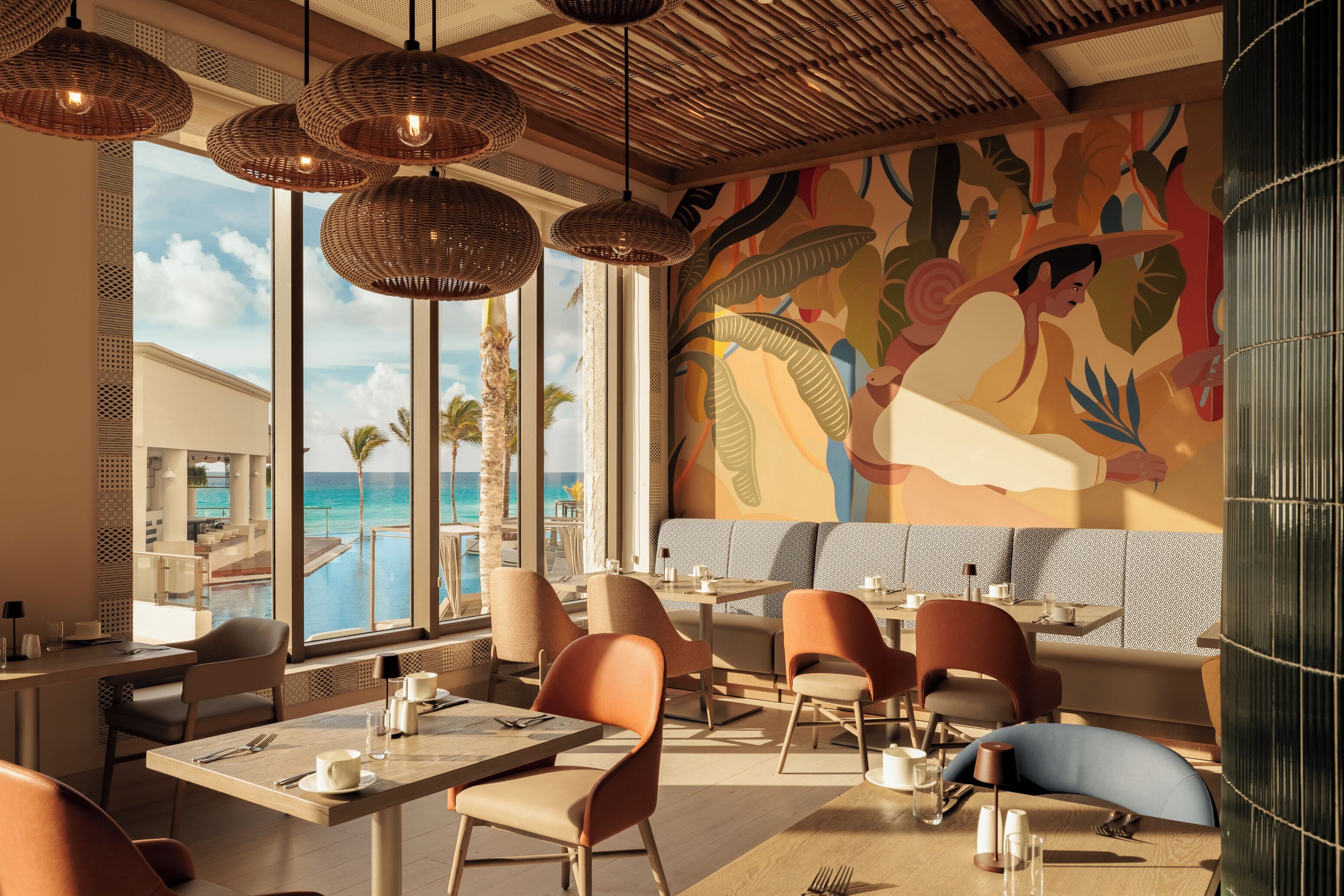 Un restaurante con vistas al océano, con lámparas colgantes de mimbre y un mural con motivos tropicales.