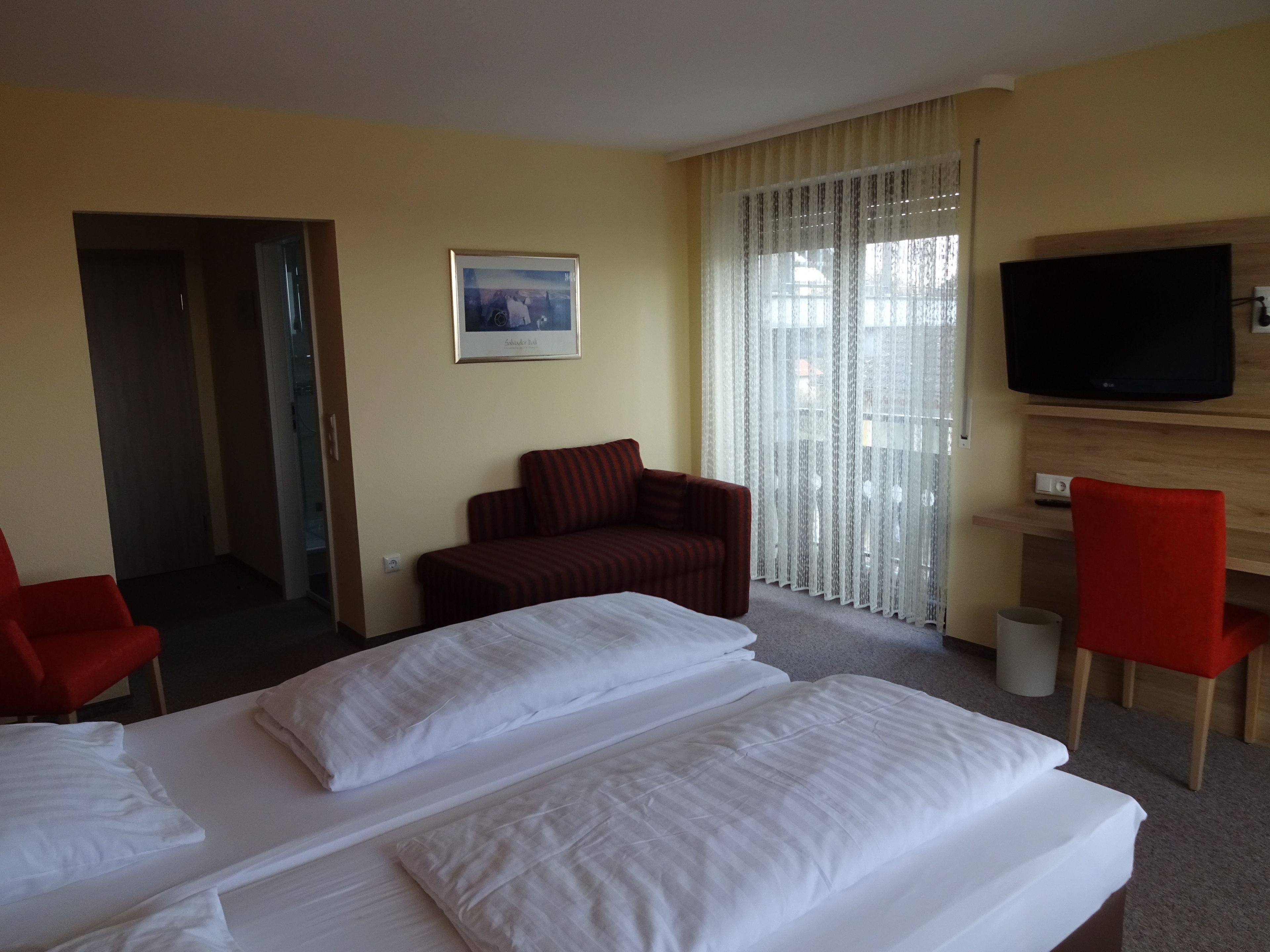 A hotel room with two beds, a red armchair, a wooden TV stand, and a large window with sheer curtains.