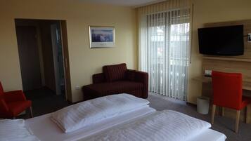 A hotel room with two beds, a red armchair, a wooden TV stand, and a large window with sheer curtains.