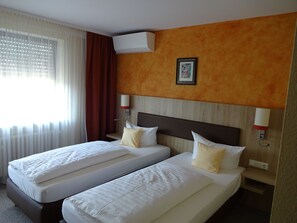Une chambre d’hôtel avec deux lits, une fenêtre avec des rideaux légers, un appareil de climatisation et un tableau encadré au mur.