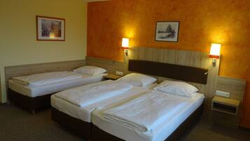 A hotel room with two single beds and a larger bed, all neatly made. There are two wall-mounted lamps providing light.