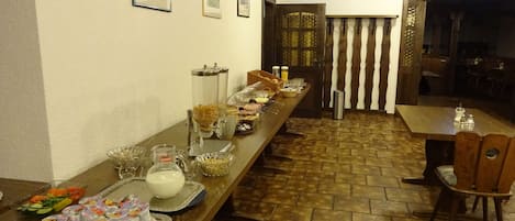 Una mesa larga con varios alimentos y bebidas en un bufé, con piso de baldosas y sillas de madera.