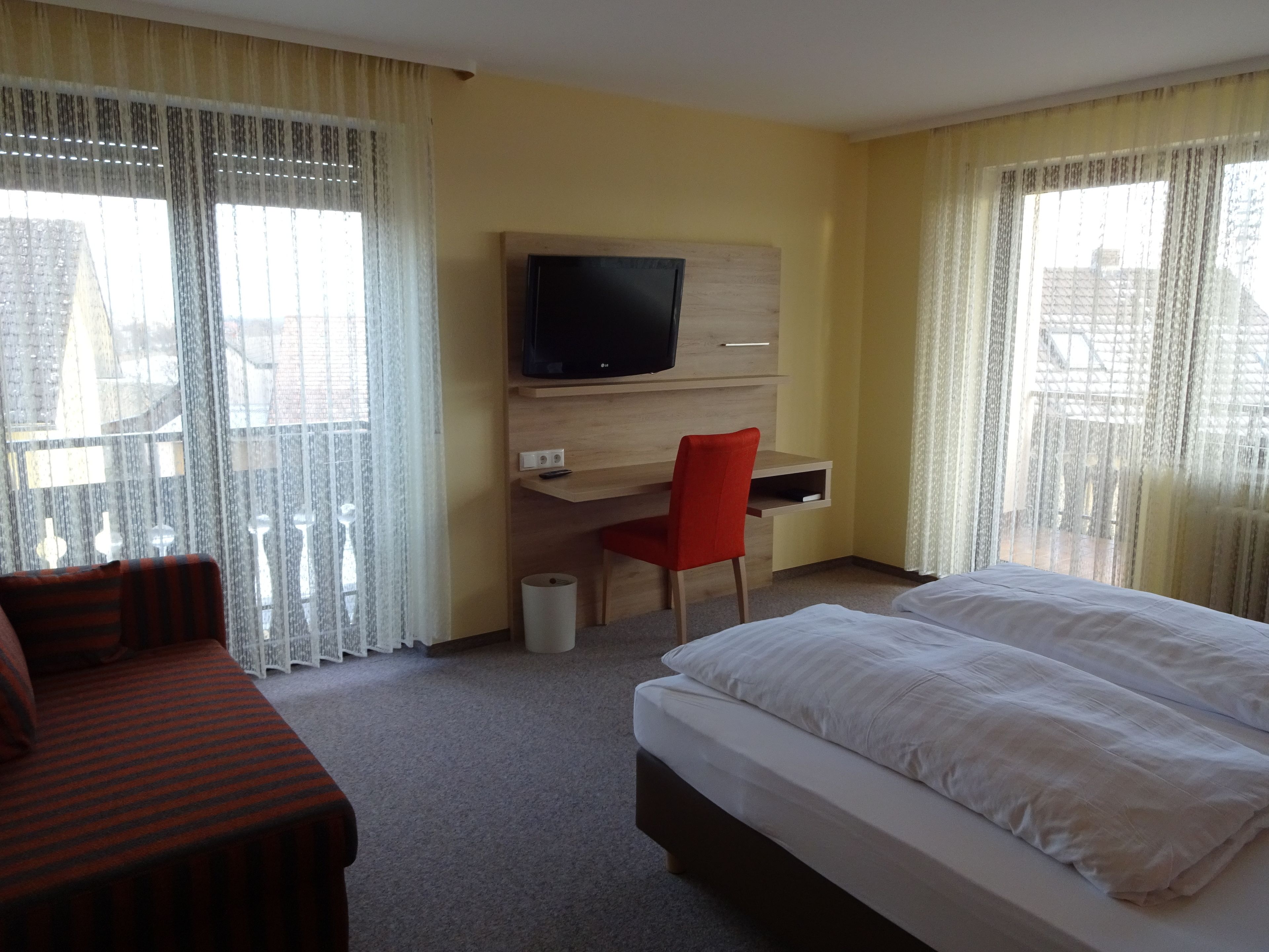 A hotel room with a bed, a desk with a chair, a television mounted on the wall, and a window with curtains.