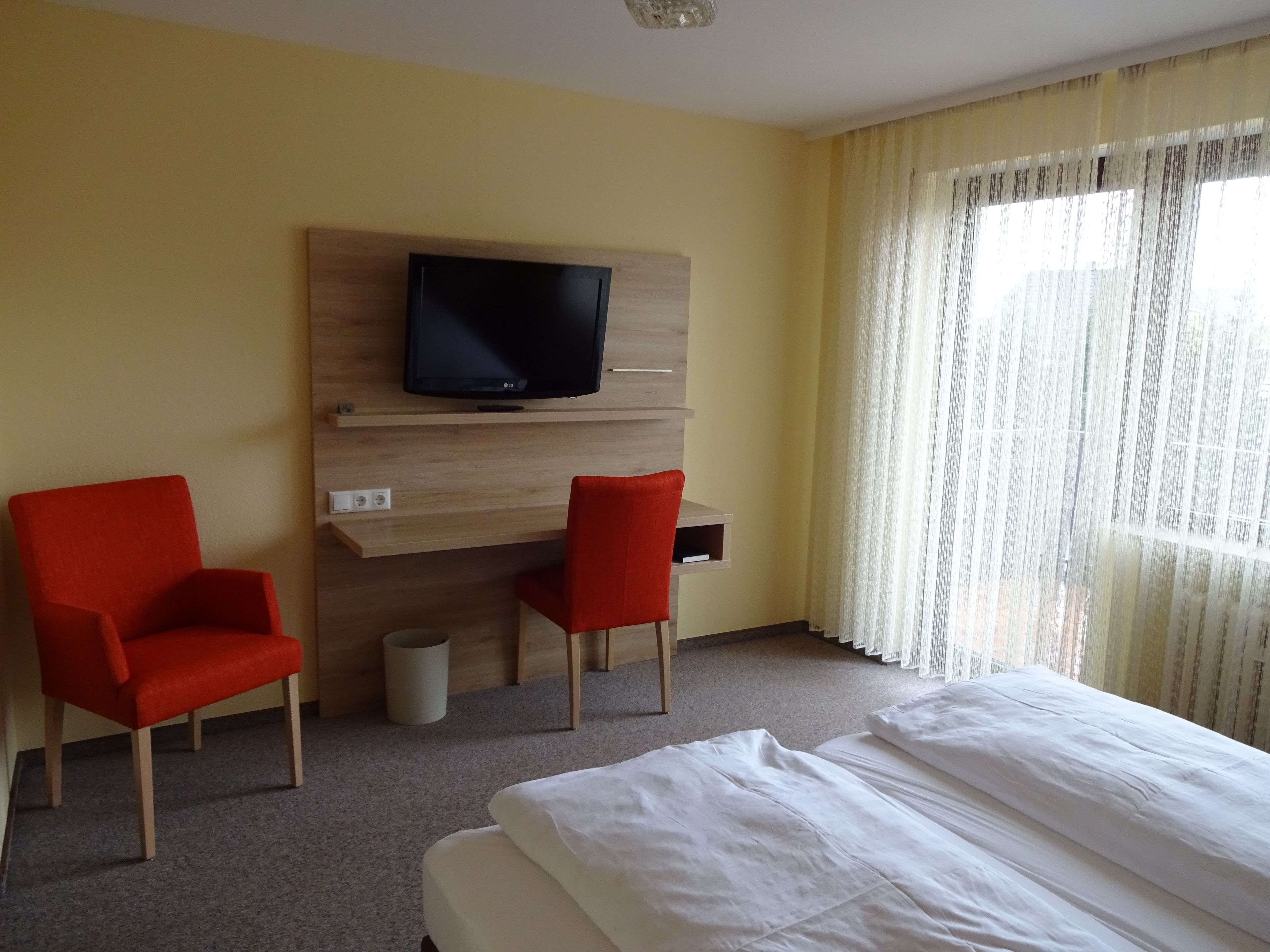 Una habitación de hotel con una cama, dos sillas rojas, un televisor sobre una estantería de madera y una ventana con cortinas translúcidas.