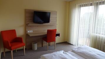 Una habitación de hotel con una cama, dos sillas rojas, un televisor sobre una estantería de madera y una ventana con cortinas translúcidas.