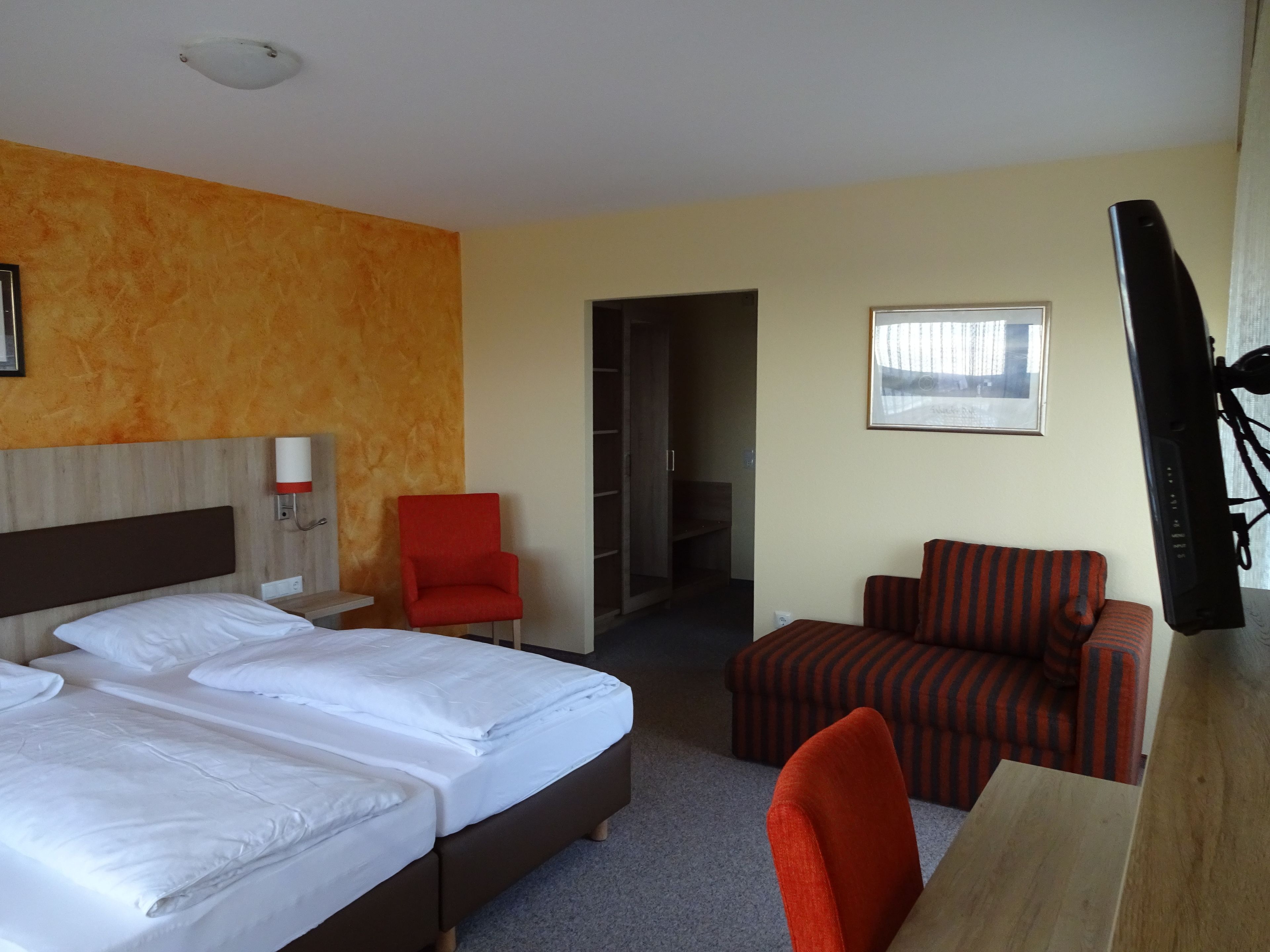 A hotel room with a bed, a red armchair, a striped sofa, a TV, and a framed picture on the wall.
