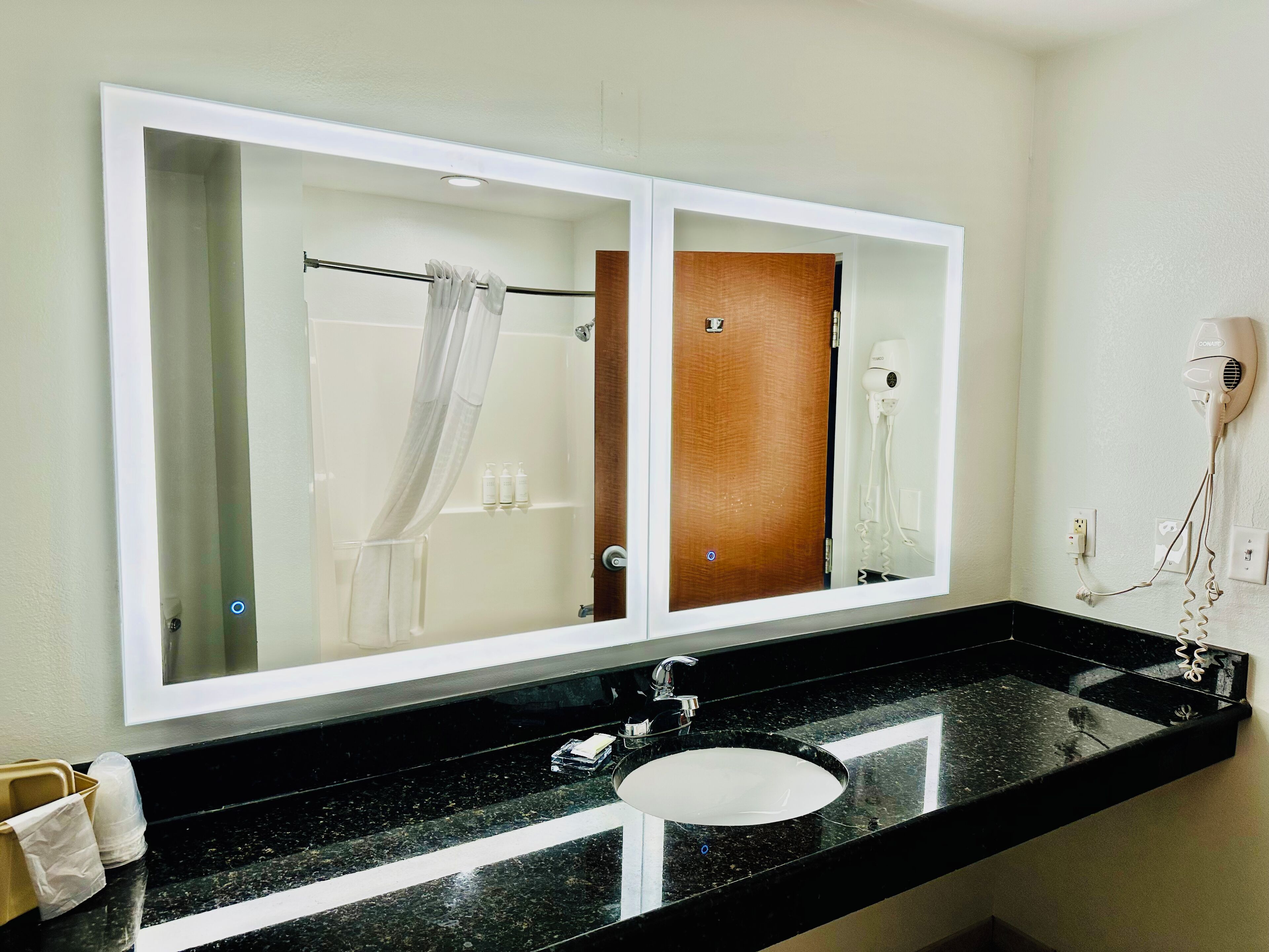 Un bagno con un grande specchio, un piano di lavoro nero, un lavandino bianco, un porta asciugamani e un asciugacapelli.