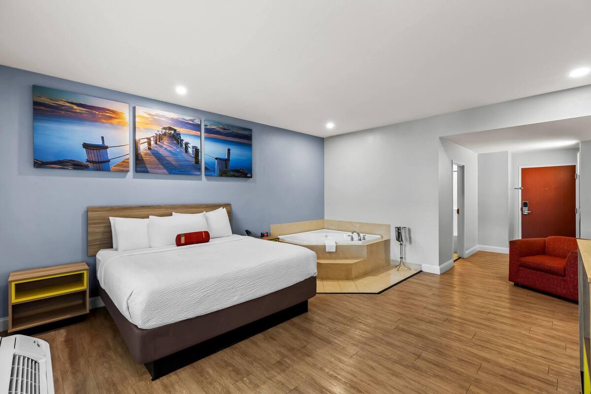 Una habitación de hotel moderna con una cama grande, una zona de estar con una silla roja, una bañera y dos cuadros con paisajes de atardecer en la pared.