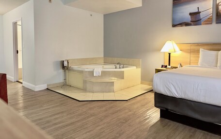 A hotel room with a large bathtub, a bed, a nightstand with a lamp, and a painting of a sunset over the ocean.
