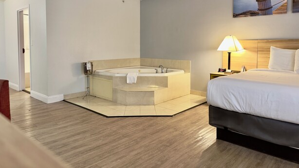 A hotel room with a large bathtub, a bed, a nightstand with a lamp, and a painting of a sunset over the ocean.