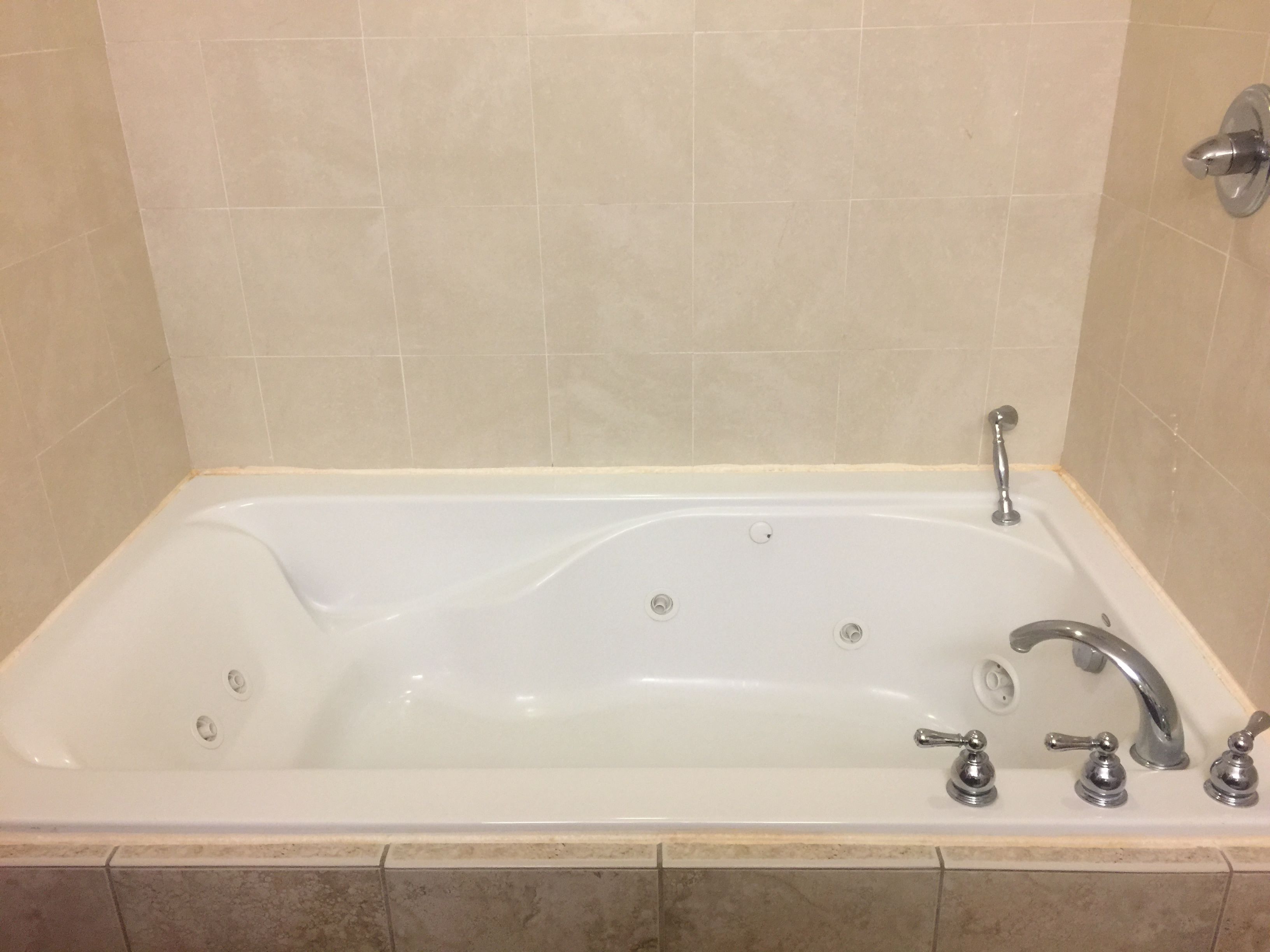 A rectangular bathtub with a single faucet and three separate controls, set against beige tiled walls.