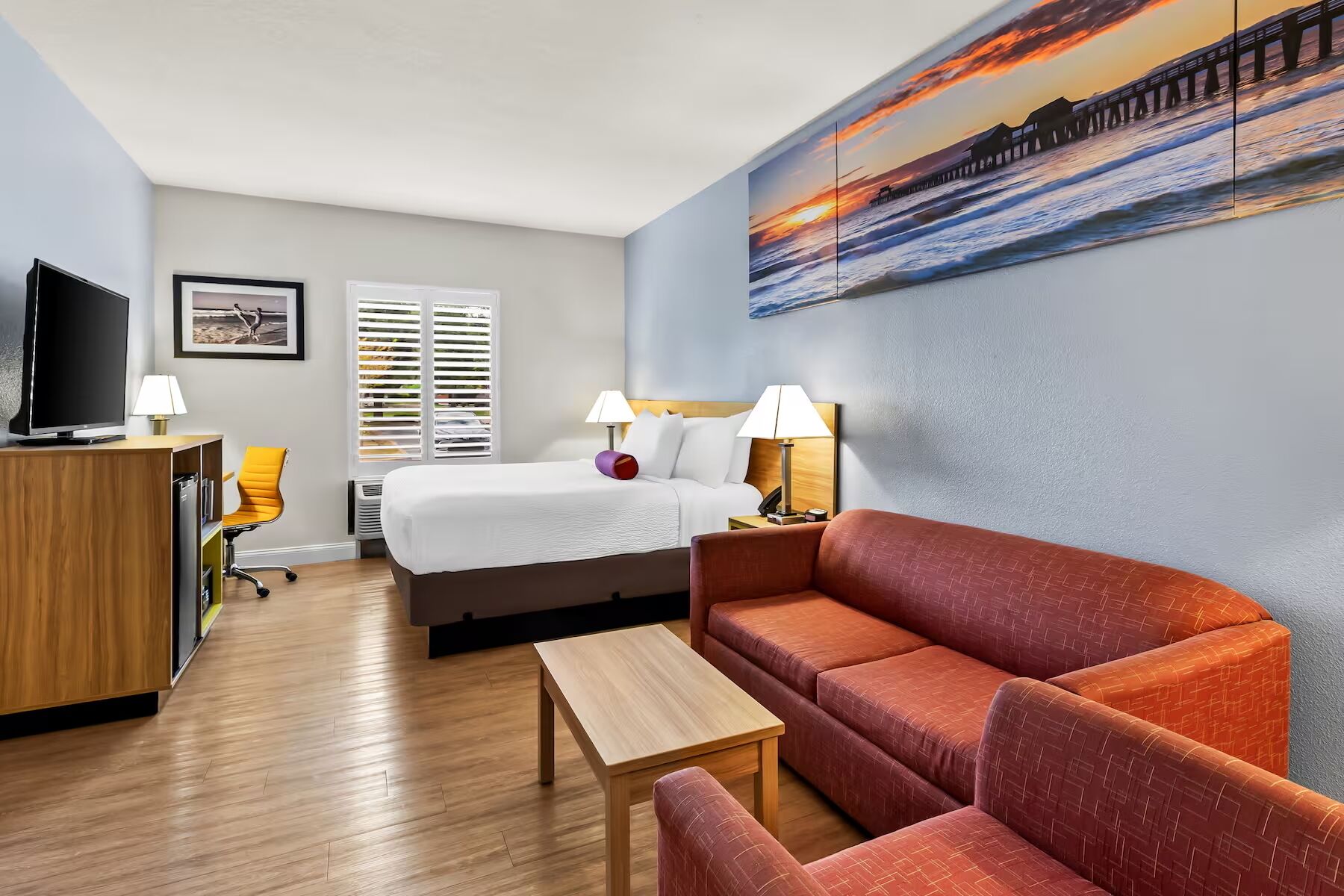 Una habitación de hotel con cama, sofá, escritorio, televisor y un cuadro de un atardecer sobre el océano.
