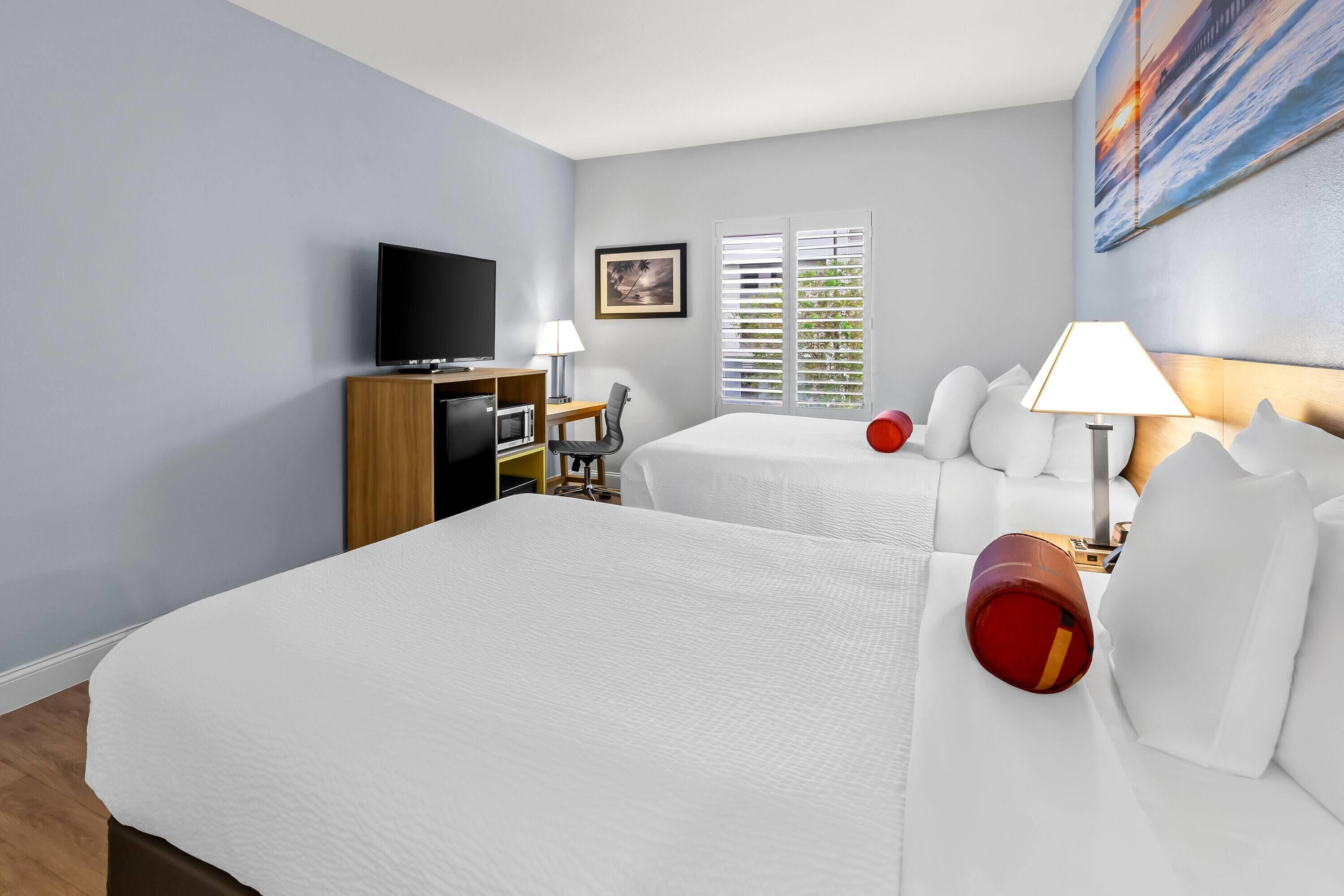 Habitación de hotel con dos camas, televisión, escritorio con silla y ventana con persianas.