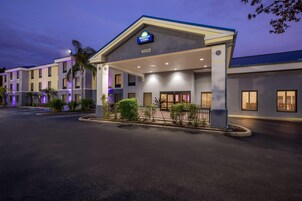 Entrada de un hotel con un letrero que dice "Days Inn Suites 4502", que presenta un acceso cubierto, un estacionamiento y una zona ajardinada con plantas pequeñas y una palmera.