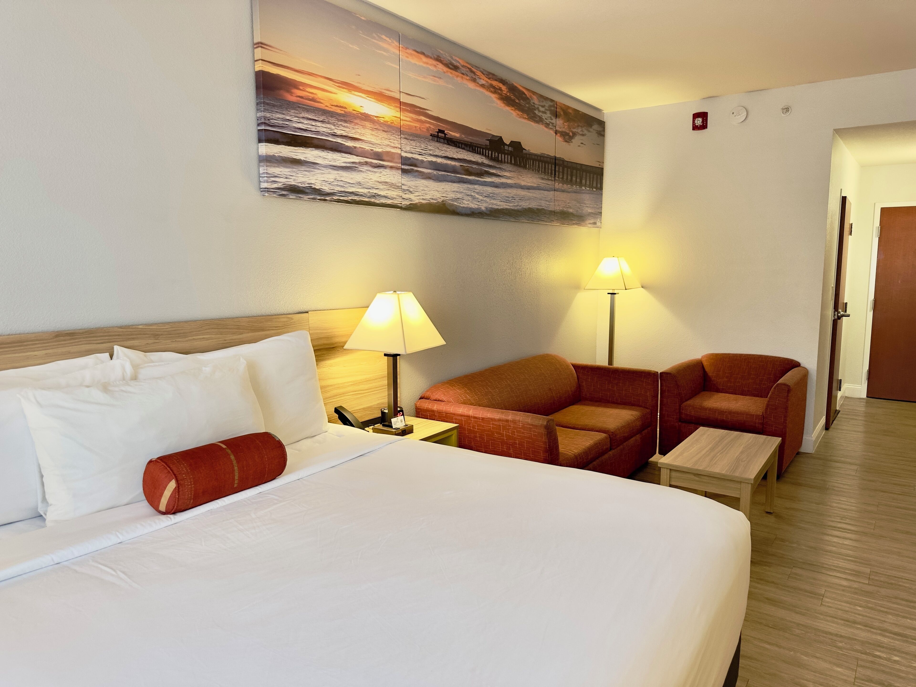 A hotel room with a large bed, two armchairs, a small table, and a painting of a sunset over the ocean.