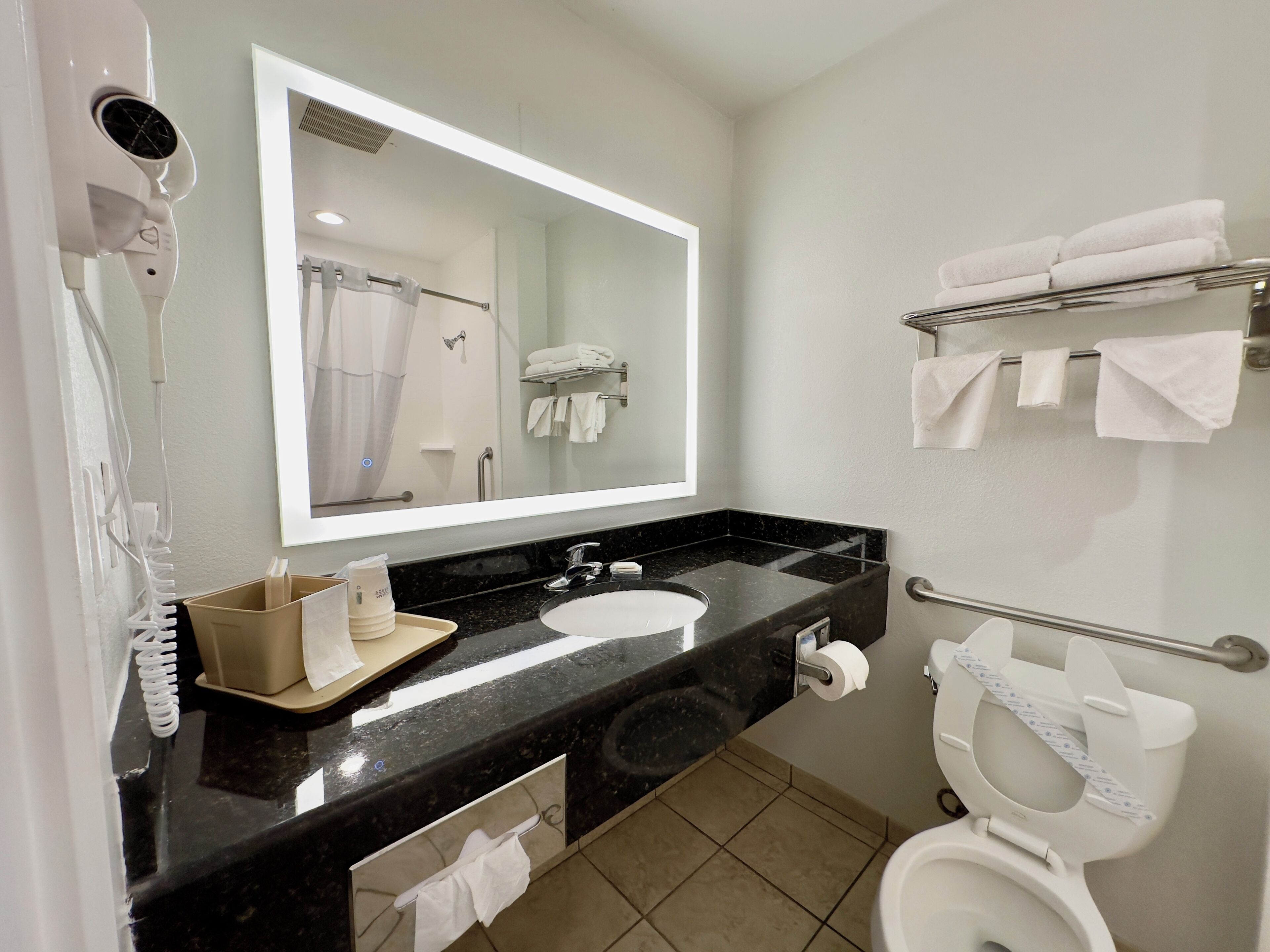 A modern bathroom with a black marble sink, a large mirror, a hairdryer, and a toilet with a towel rack.