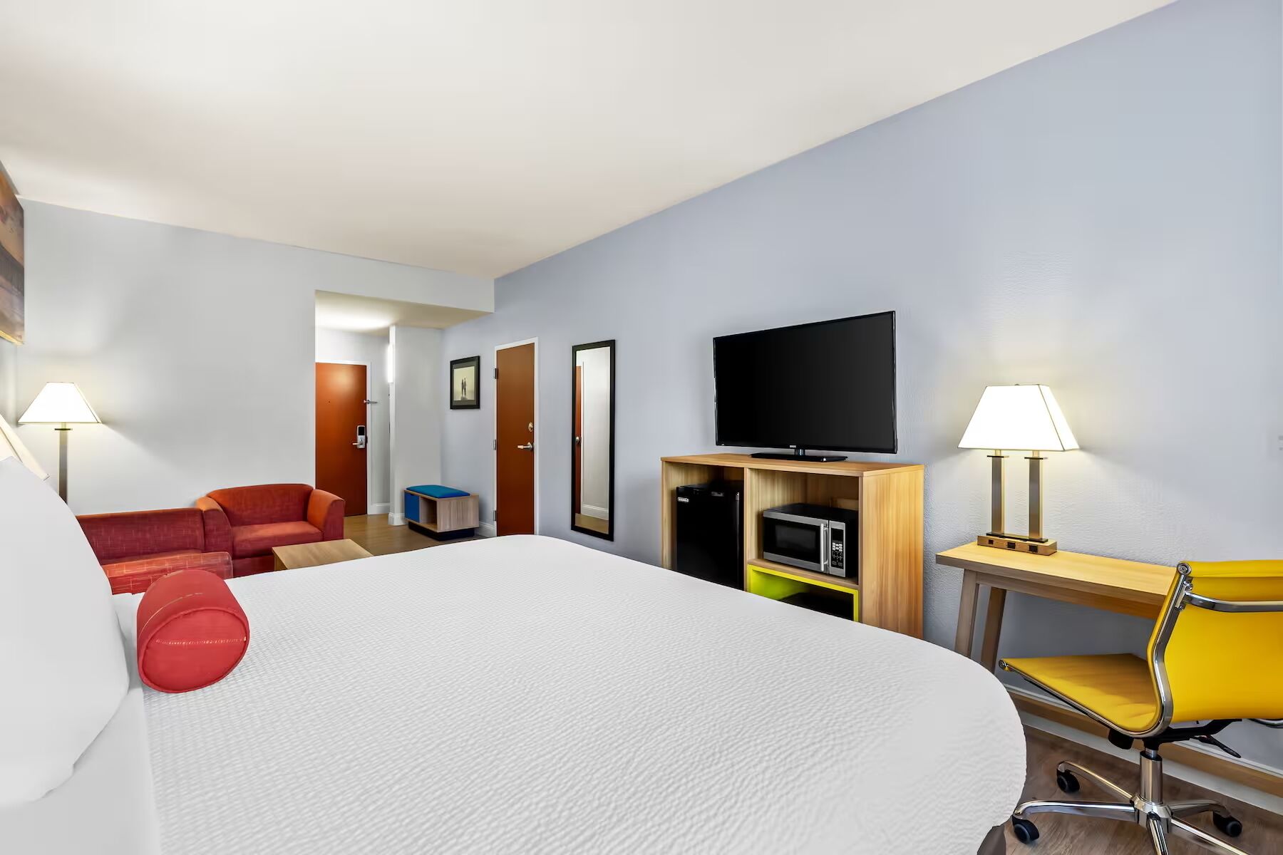 A hotel room with a bed, a desk with a chair, a television, a microwave, and a small seating area with a red couch and a yellow chair.