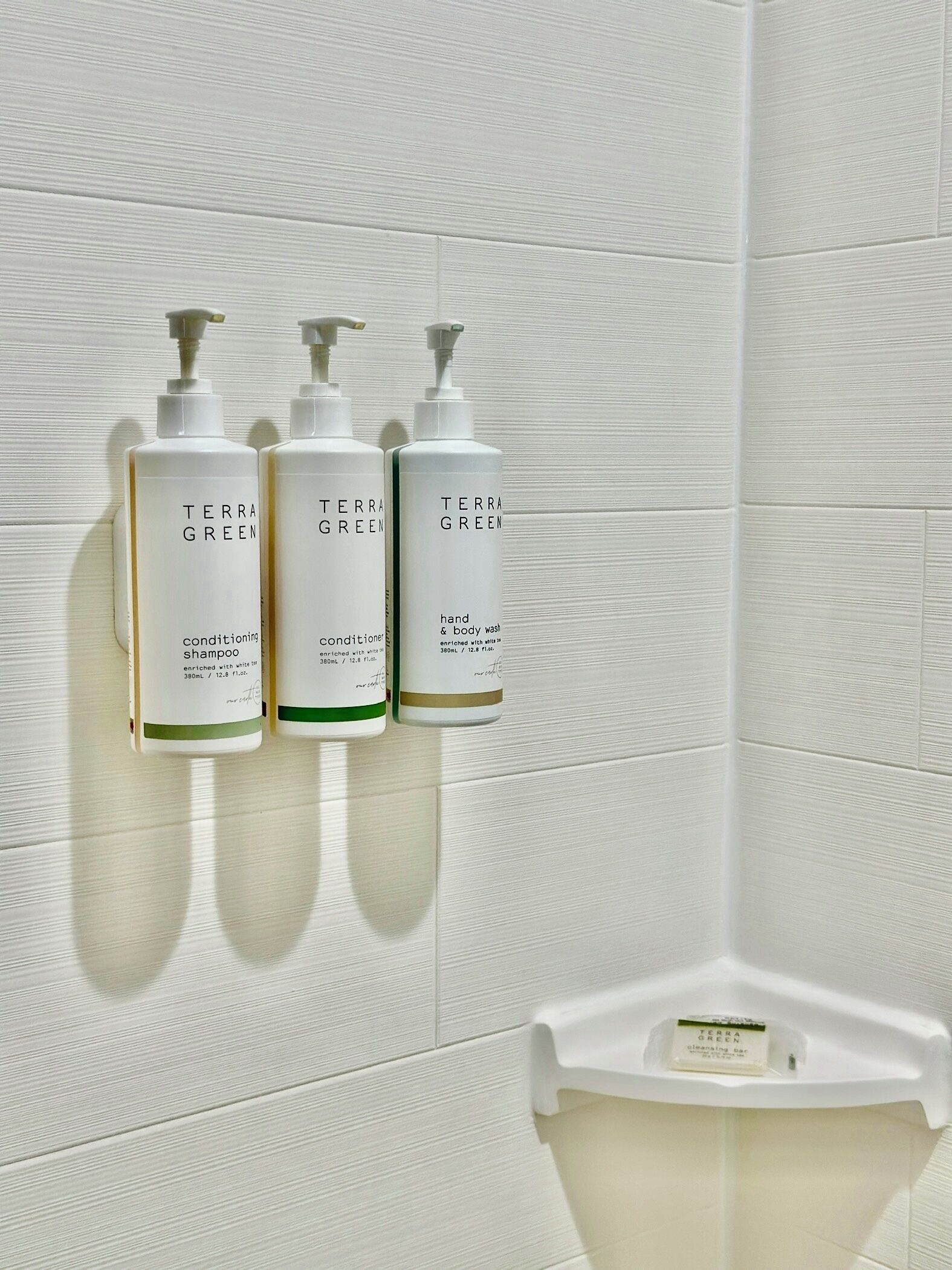 Tres botellas de jabón líquido Terra Green para manos y cuerpo sobre una pared de baldosas blancas.