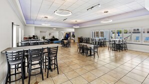 A spacious dining area with multiple tables and chairs, a kitchen in the background, and large windows providing natural light.