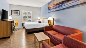 A hotel room with a bed, a sofa, a desk, a TV, and a painting of a sunset over the ocean.