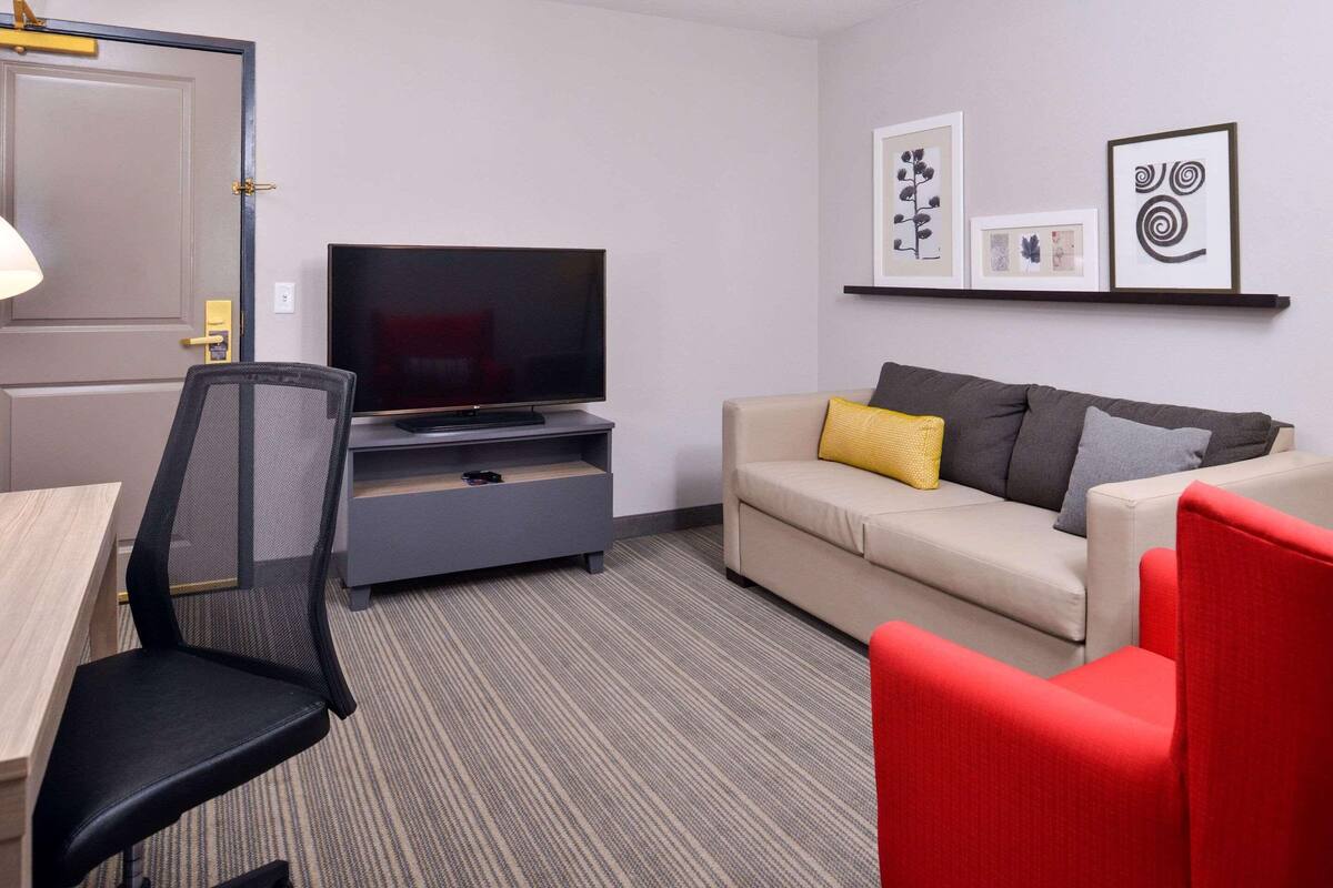Une chambre d’hôtel moderne équipée d’un téléviseur à écran plat posé sur un meuble, d’un canapé avec des coussins, d’un fauteuil rouge, d’un bureau avec une chaise et d’une porte dotée d’une poignée dorée.