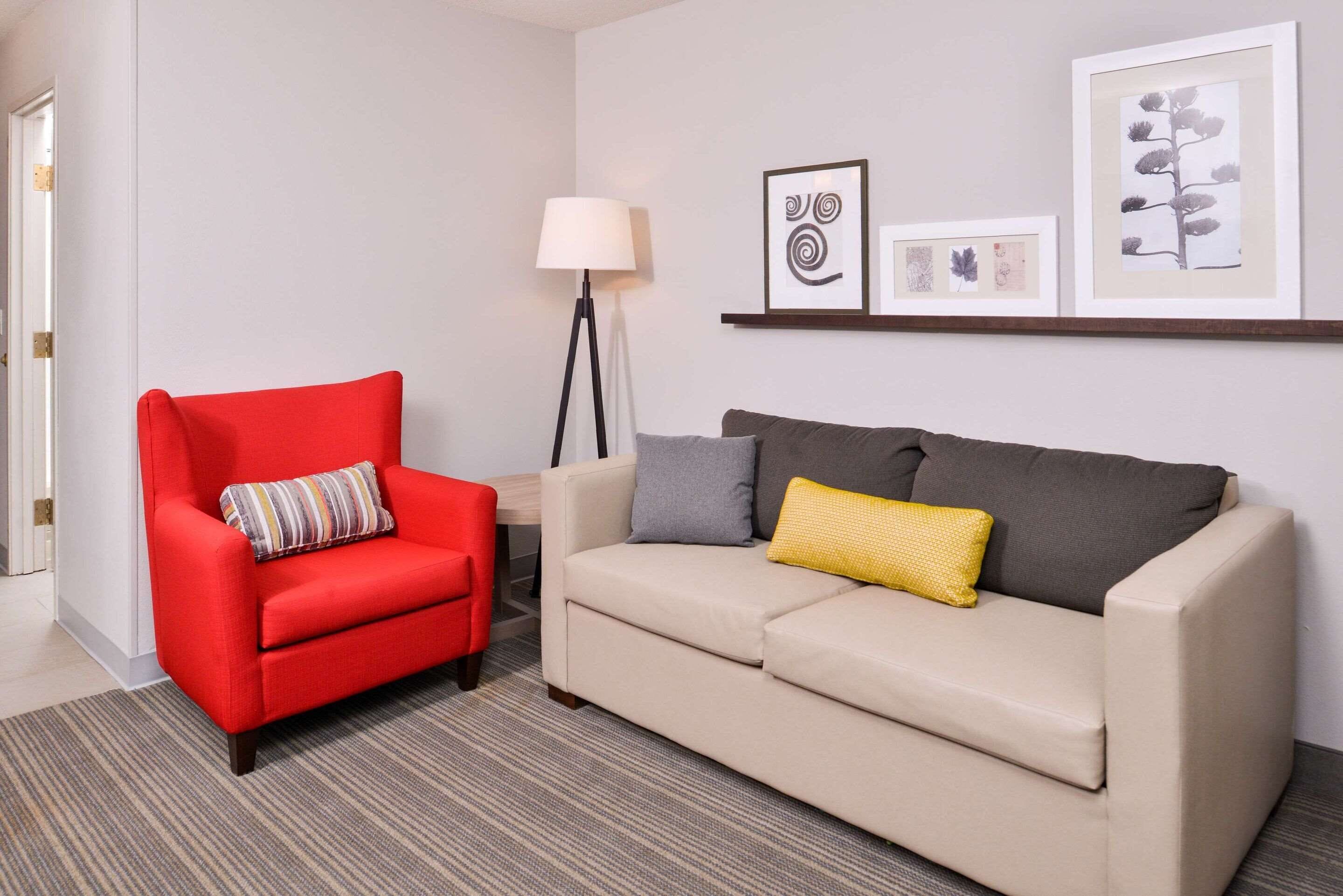 A room with a red armchair, a beige sofa, a floor lamp, and framed botanical prints on the wall.