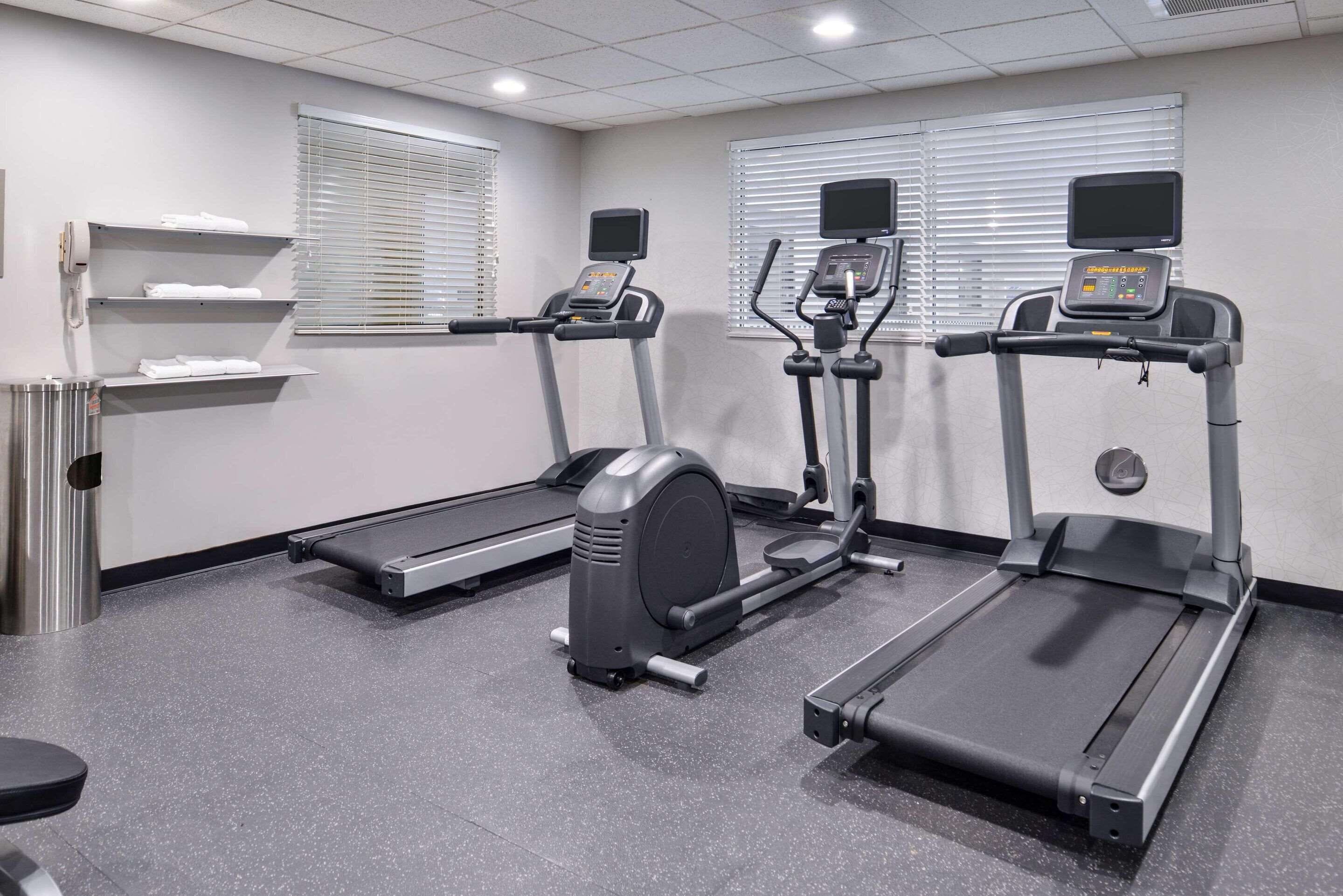 A gym with treadmills, an elliptical, and a stationary bike. There are windows with blinds, a trash can, and a shelf with towels.