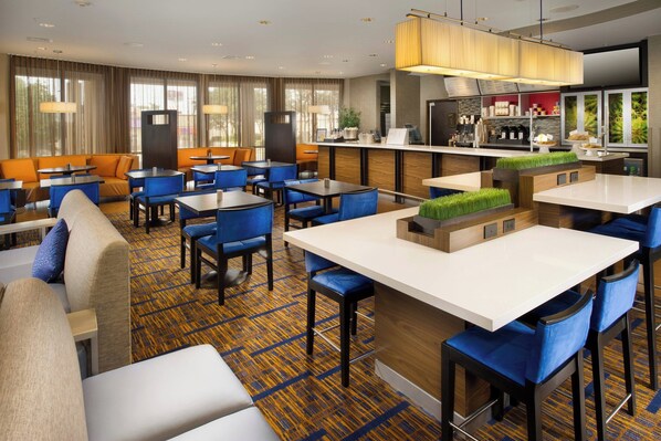 A hotel lobby with a mix of seating areas, including sofas and tables, a bar area, and a kitchen visible in the background.