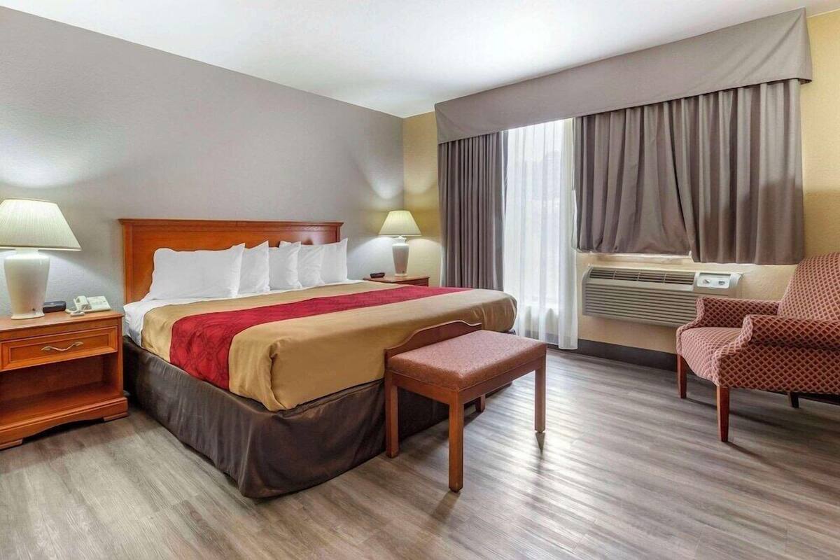 A hotel room with a bed, bedside tables, a chair, an air conditioning unit, and a window with curtains.