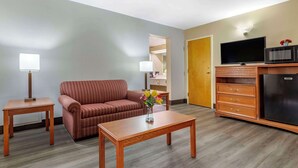 A hotel room with a sofa, a wooden coffee table, a TV on a cabinet, and a lamp on a side table. - Econo Lodge & Suites Southern Pines - Pinehurst (Southern Pines)