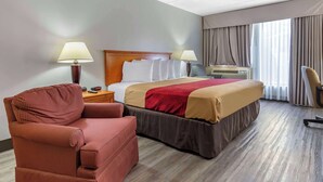 A hotel room with a bed, a red armchair, a desk with a lamp, and a window with curtains. - Econo Lodge & Suites Southern Pines - Pinehurst (Southern Pines)
