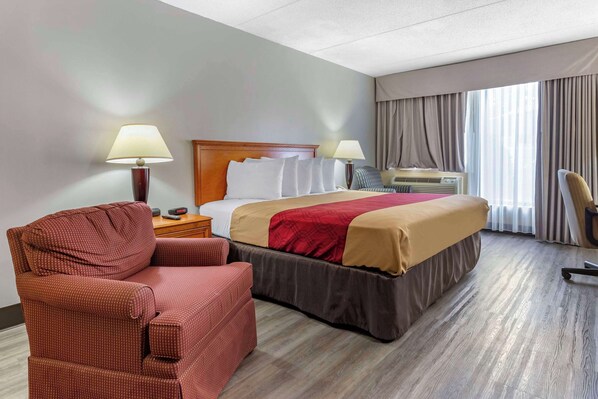 A hotel room with a bed, a red armchair, a desk with a lamp, and a window with curtains.