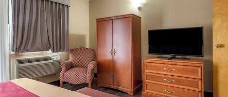 A hotel room with a bed, a TV on a wooden dresser, a wardrobe, a patterned armchair, and a window with curtains.