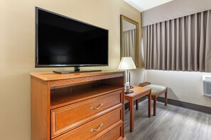 A hotel room with a flat-screen TV, a wooden dresser with drawers, a small table with a lamp, a chair, and a large window with curtains. - Econo Lodge & Suites Southern Pines - Pinehurst (Southern Pines)