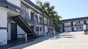 A multi-story motel with a staircase leading to the entrance, outdoor seating area, and palm trees. - Value Inn Bellflower (Bellflower)