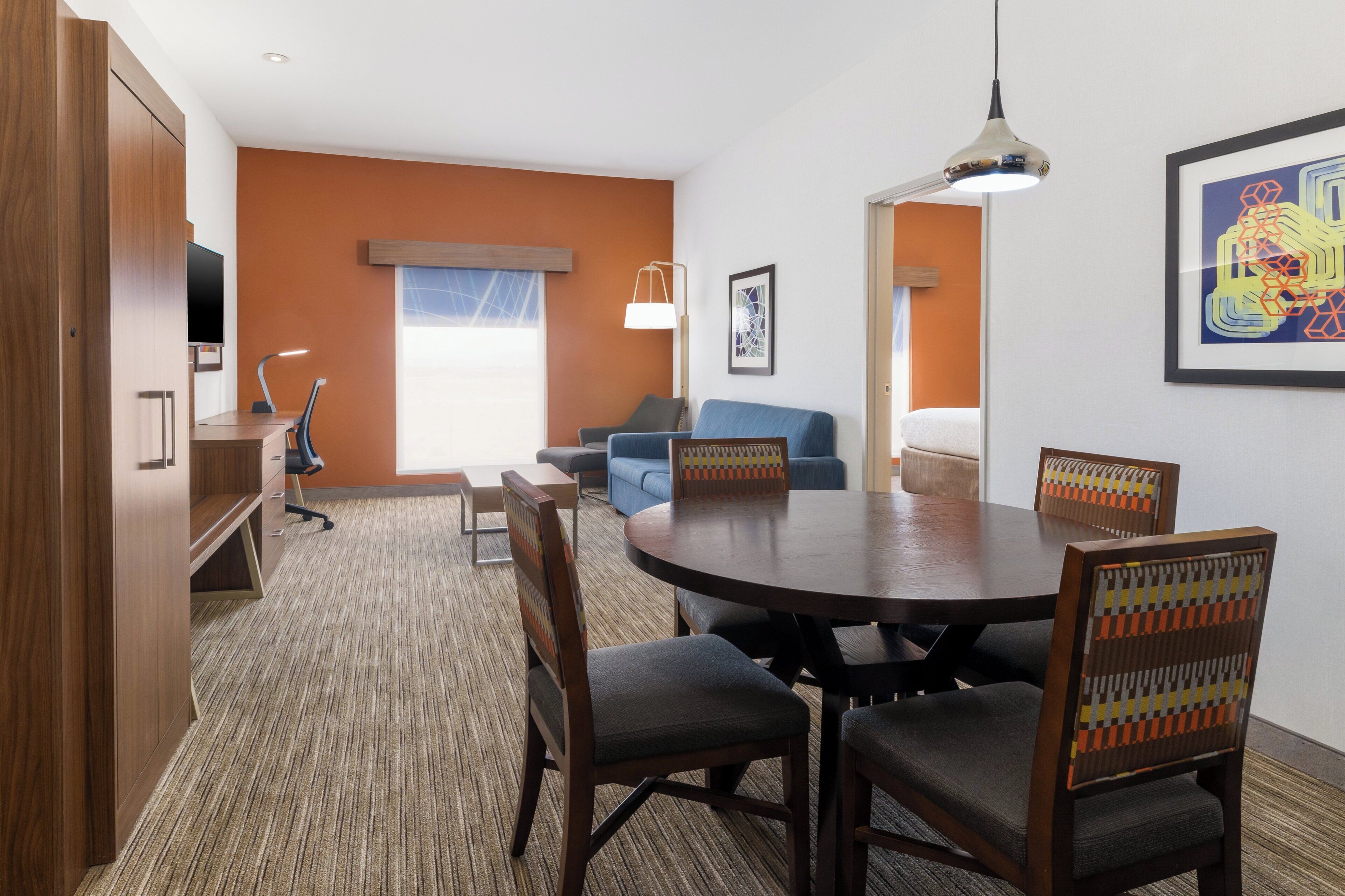 Quarto de hotel com mesa de jantar redonda, cadeiras de madeira, sofá azul, televisão e uma obra de arte emoldurada na parede.