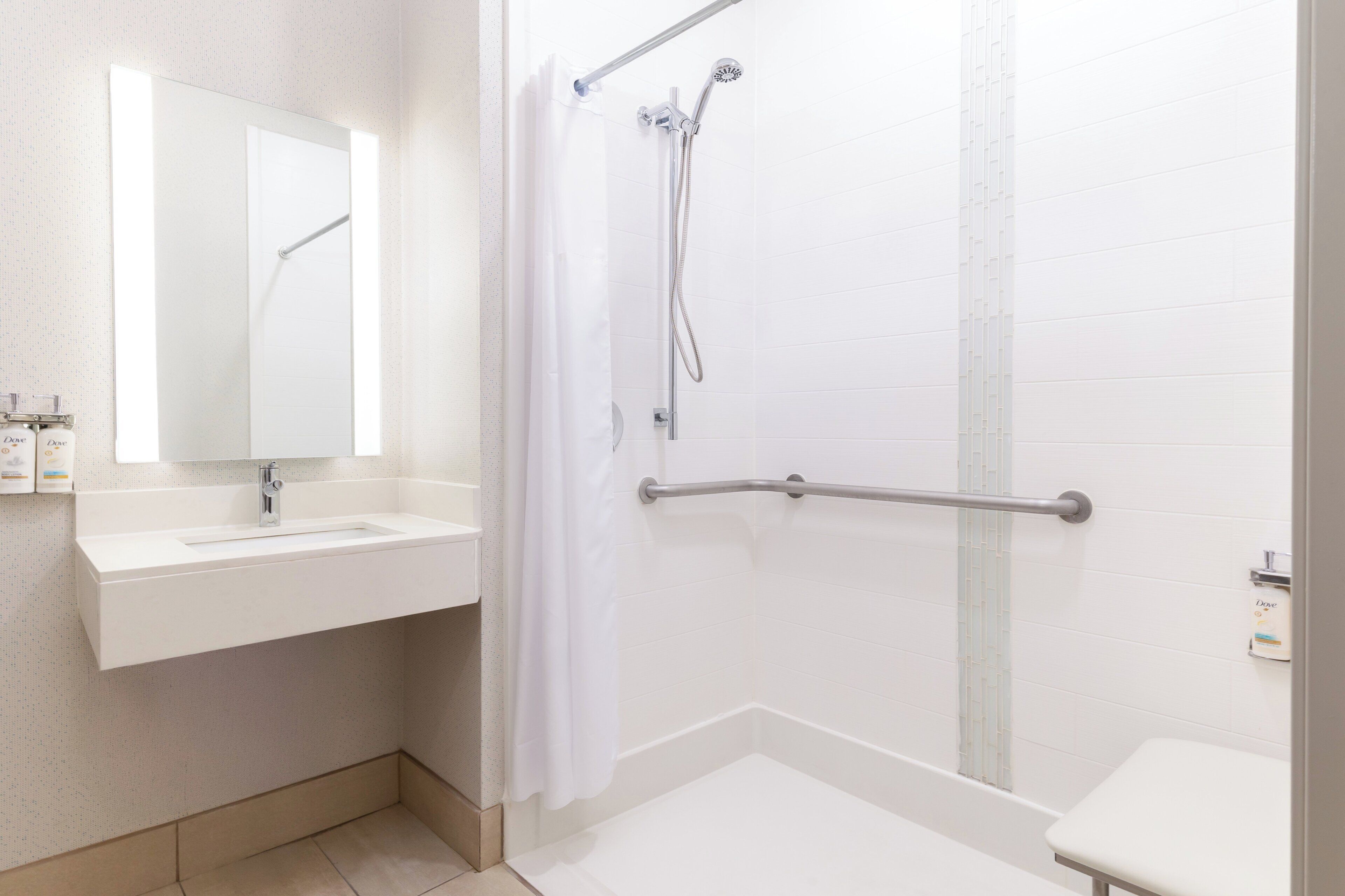 Un baño con lavamanos blanco, espejo, ducha con barras de apoyo y un inodoro.