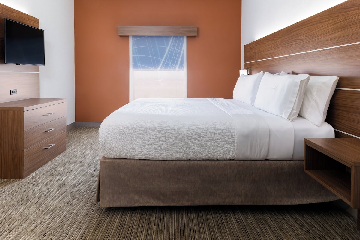 A modern hotel room with a large bed, wooden headboard, a flat-screen TV, and a window with a view of the sky.