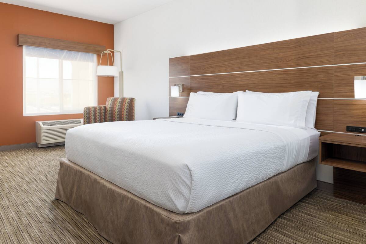 A hotel room with a large bed, a bedside table, a chair, a window with blinds, and an air conditioning unit.